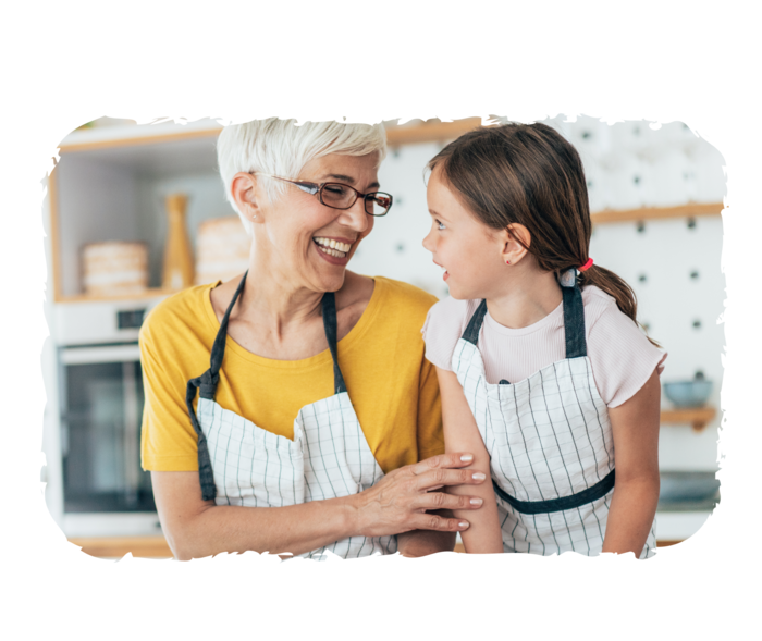 Eine Frau und ein junges Mädchen tragen Kochschürzen und lachen. Vor ihnen liegen verschiedene Lebensmittel auf einem Tisch.