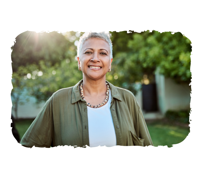 Eine ältere Frau steht in einem Garten und lächelt in die Kamera.