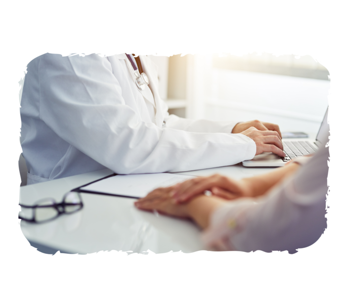 Fotoausschnitt: Zwei Personen sitzen an einem Tisch, die Arme sind dabei sichtbar. Eine Person trägt einen weißen Kittel und hat ein Stethoskop um die Schultern liegen.