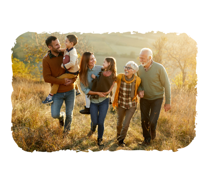 Ein Mann und eine Frau tragen jeweils ein Kind auf dem Arm, daneben ein älterer Mann und eine ältere Frau. Die Personen laufen eine Wiese entlang und lachen.