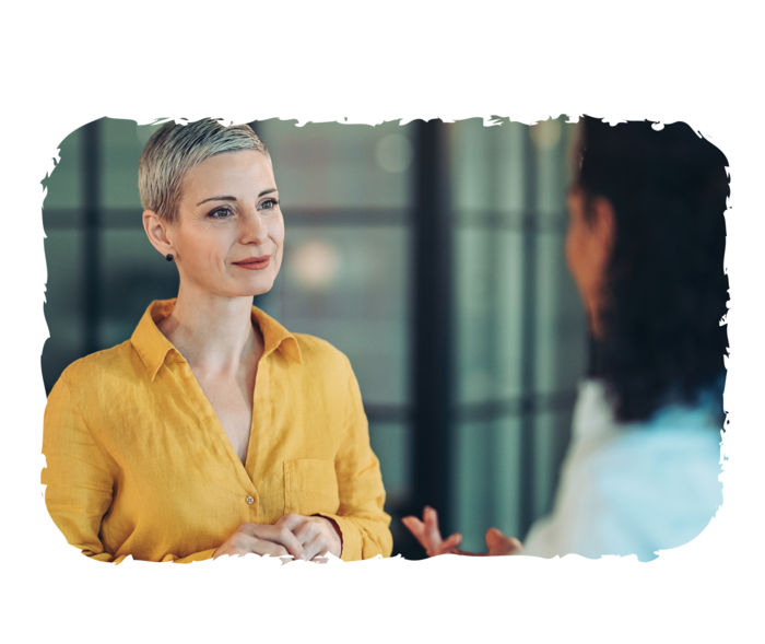 Zwei Frauen stehen sich bei einem Gespräch gegenüber: Die eine Frau ist von hinten zu sehen und spricht, die andere Frau ist von vorn zu sehen und hört zu.