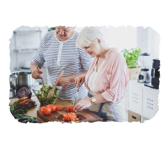 Eine Frau und ein Mann im Seniorenalter bereiten mit frischen Zutaten einen Salat zu: Die Frau schneidet Tomaten, der Mann mischt in einer Schüssel die Salatblätter.