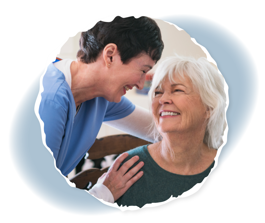 Eine Frau in Pflegekleidung und eine ältere Frau schauen sich an und lächeln.