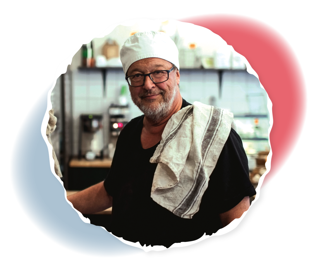 Man mit Kochmütze, grauem Bart und Brille lächelt in die Kamera und hat ein Geschirrtuch über die Schulter geworfen. 