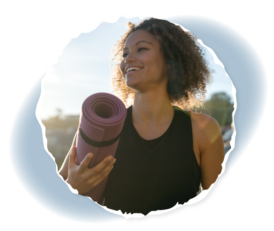 Selbstfürsorge für Angehörige: Auszeit nehmen Eine lächelnde Frau im Freien trägt eine Sportmatte unter ihrem Arm.