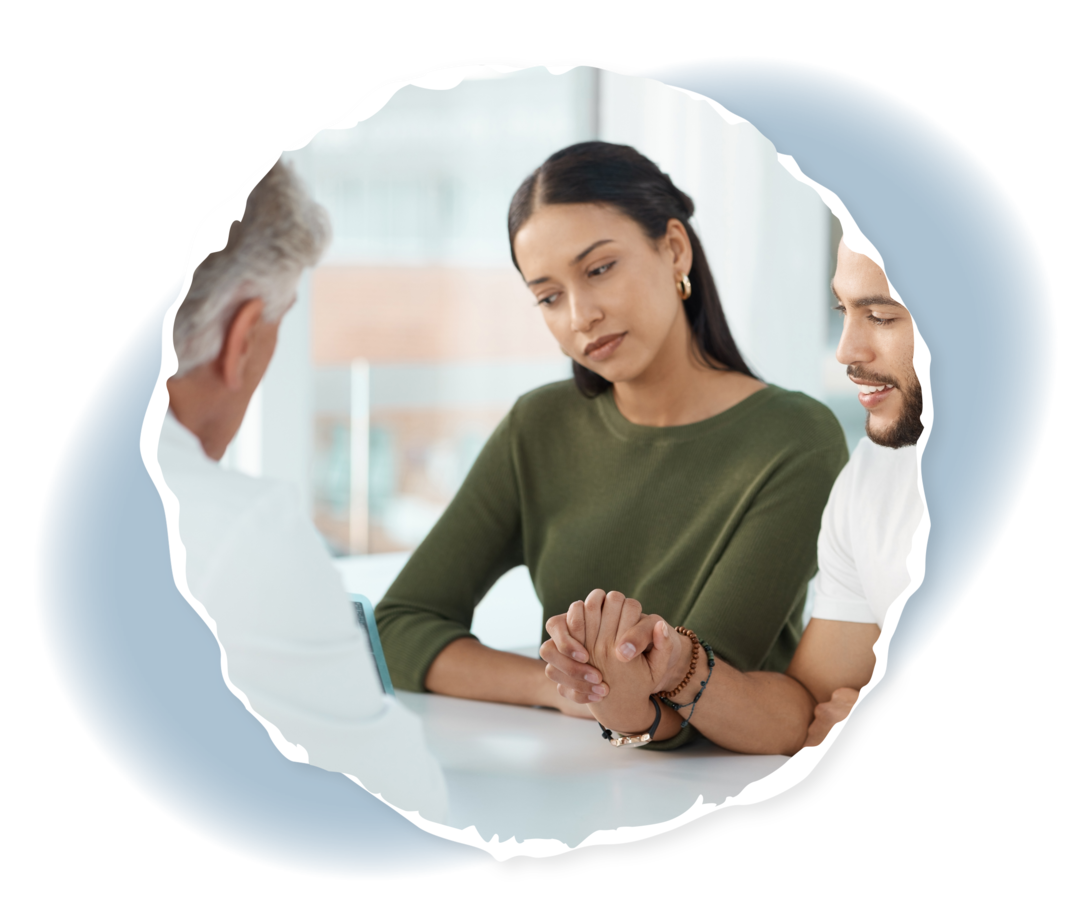 Ein Mann und eine Frau halten ihre Hände und sitzen gemeinsam mit einem Arzt an einem Tisch.