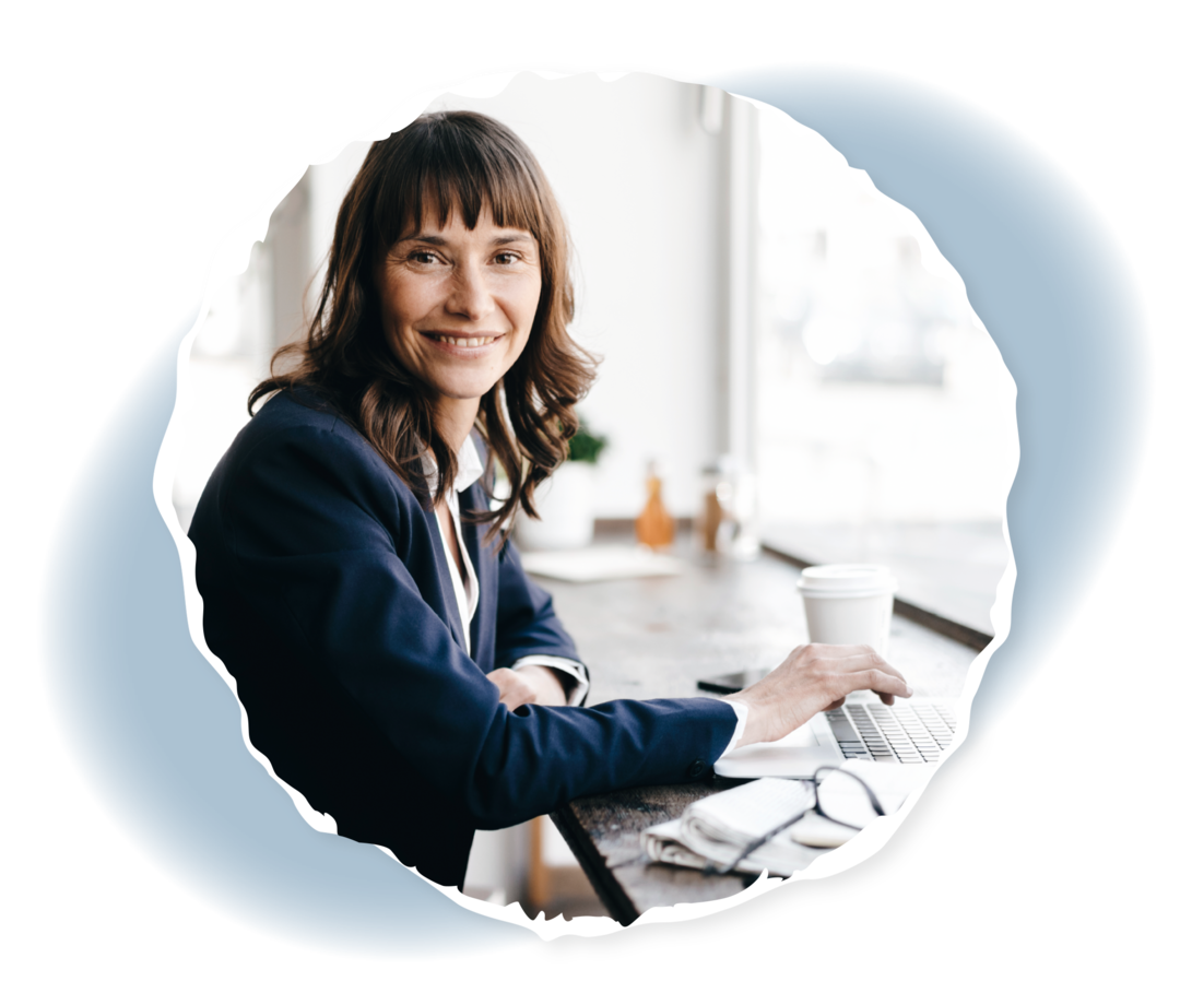 Eine Frau sitzt an einem Tisch und lächelt in die Kamera. Vor ihr ein Laptop.