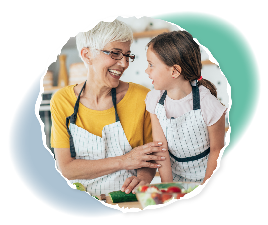 Eine Frau und ein junges Mädchen tragen Kochschürzen und lachen. Vor ihnen liegen verschiedene Lebensmittel auf einem Tisch.