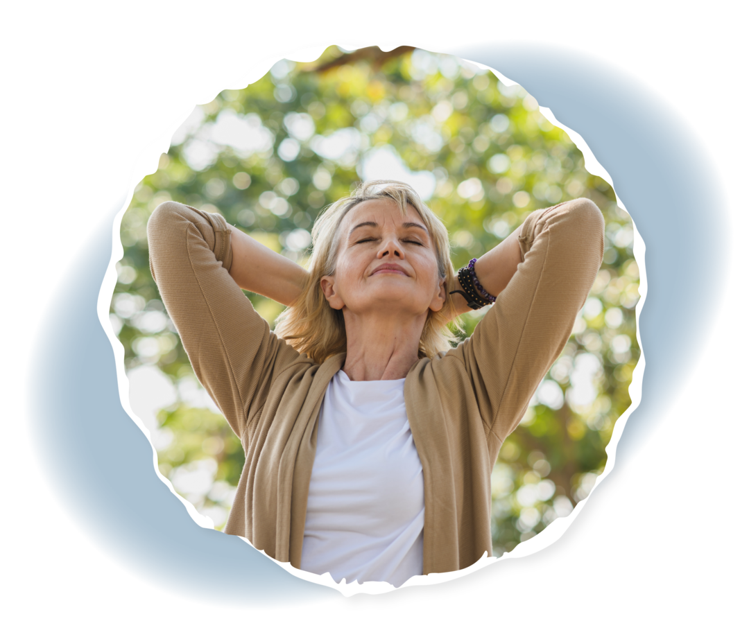 Eine ältere Frau steht mit verschlossenen Augen vor einem Baum und verschränkt die Arme hinter dem Kopf.