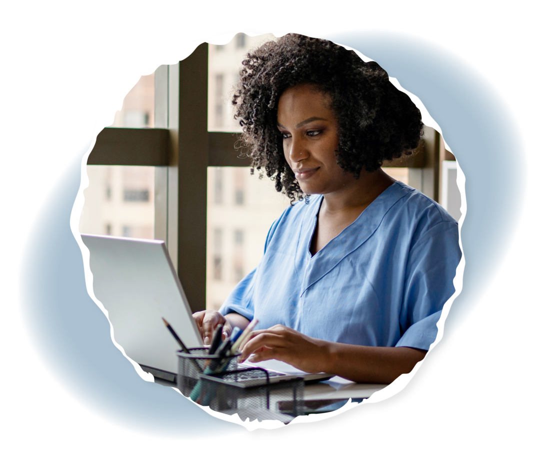 Eine Frau in Pflegekleidung sitzt an einem Schreibtisch und schaut auf einen Laptop.