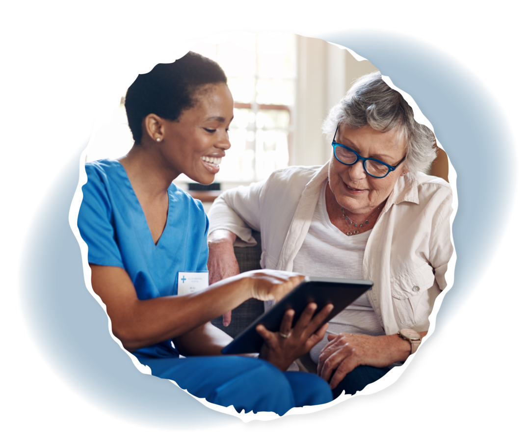 Eine Frau in Pflegekleidung hält einen Tablet-Computer. Neben ihr eine ältere Frau.