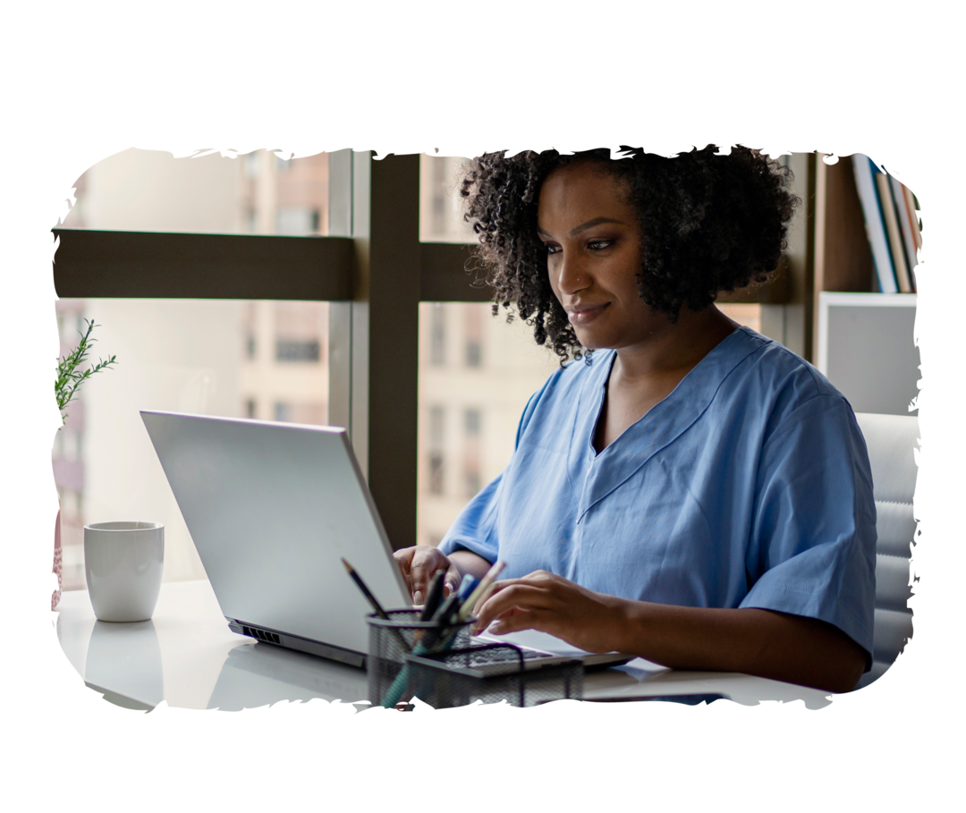 Eine Frau in Pflegekleidung sitzt an einem Schreibtisch und schaut auf einen Laptop.