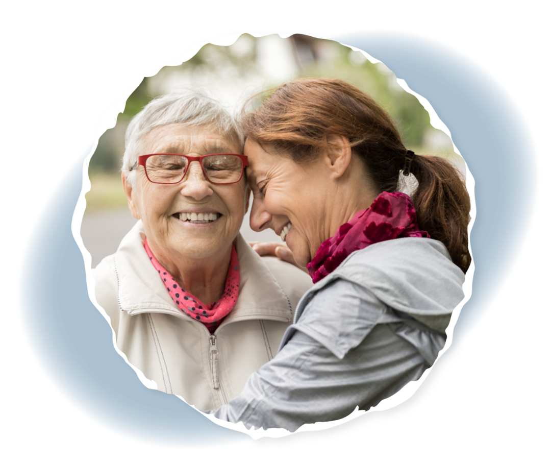 Eine Frau umarmt eine ältere Frau und lehnt ihre Stirn gegen deren Schläfe. Beide haben die Augen geschlossen und lächeln.