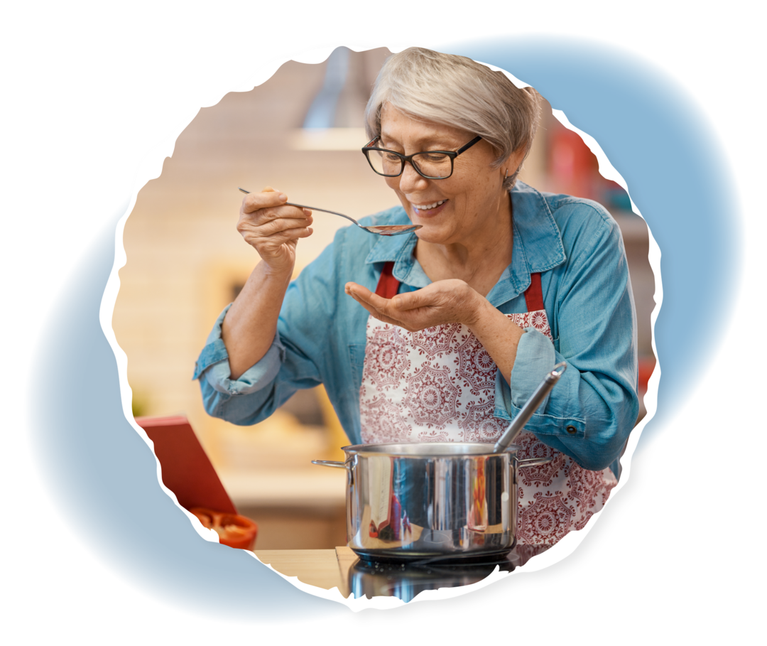 Eine ältere Frau steht am Herd und führt einen Löffel zum Mund.