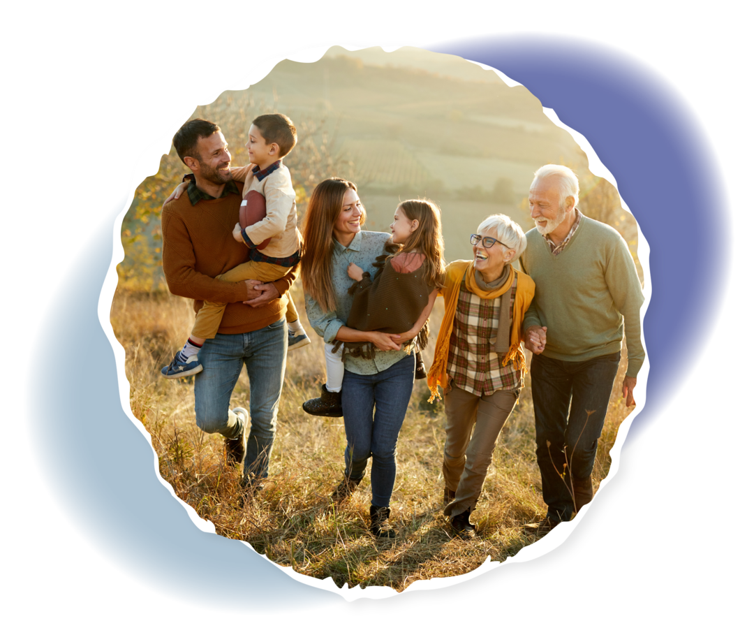 Ein Mann und eine Frau tragen jeweils ein Kind auf dem Arm, daneben ein älterer Mann und eine ältere Frau. Die Personen laufen eine Wiese entlang und lachen.