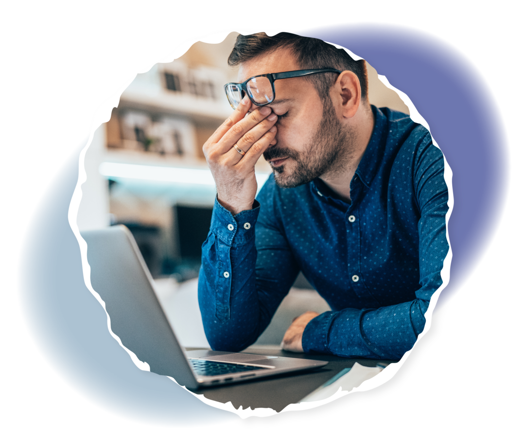 Ein bärtiger Mann in einem blauen Hemd sitzt mit geschlossenen Augen vor einem Notebook und reibt seine Nasenwurzel. Seine Brille ist hochgeschoben.