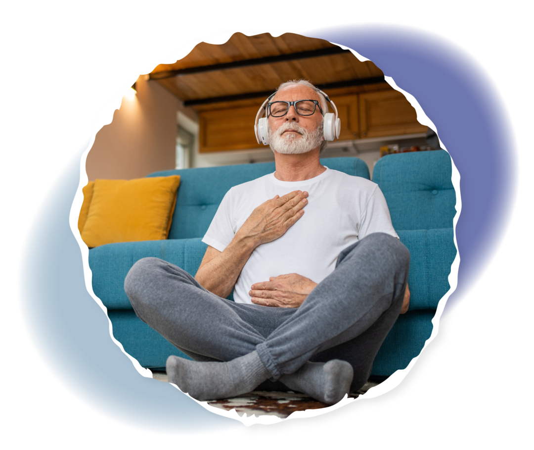 Ein Mann mit grauen Haaren lehnt im Schneidersitz an ein Sofa. Er hat die Augen geschlossen und trägt einen Kopfhörer.