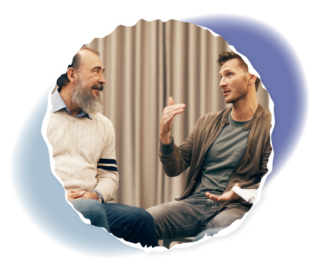 Zwei Männer sitzen vor einem Vorhang in einem Stuhlkreis und unterhalten sich.