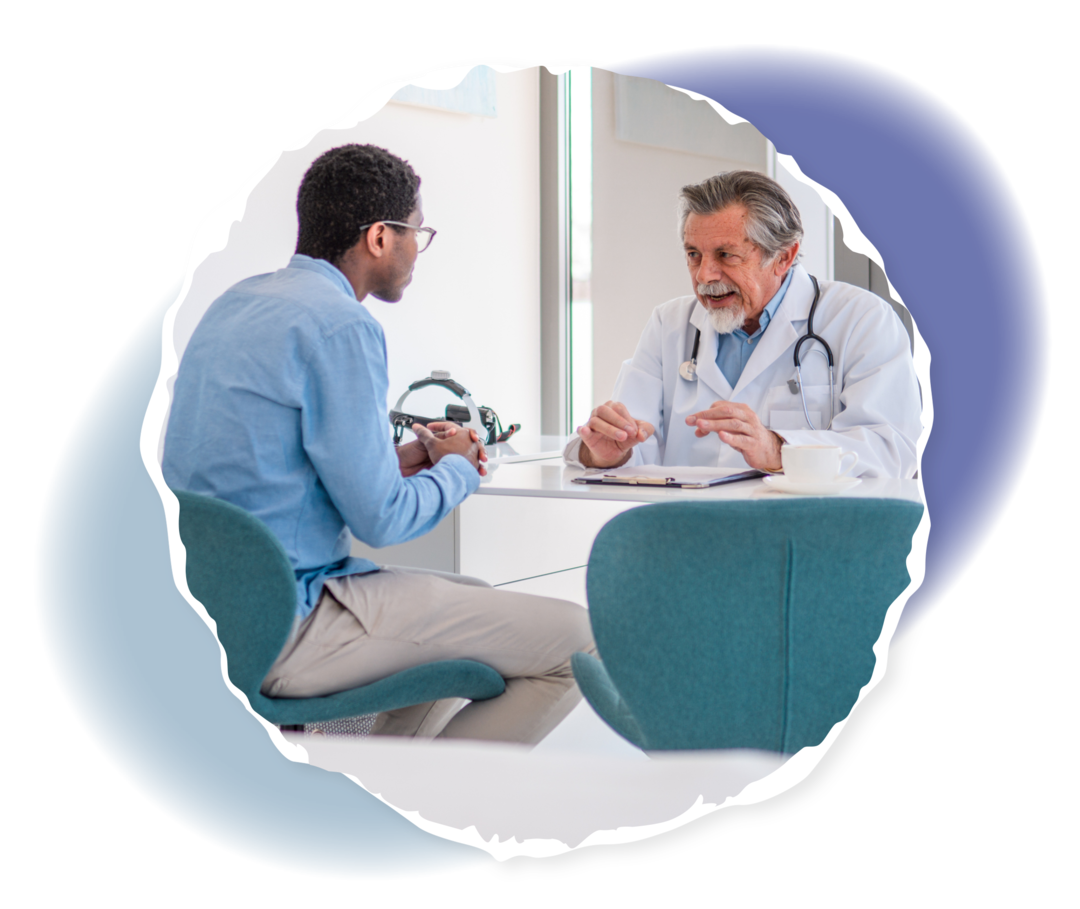 Mann mit weißem Kittel sitzt Mann an einem Tisch gegenüber. 