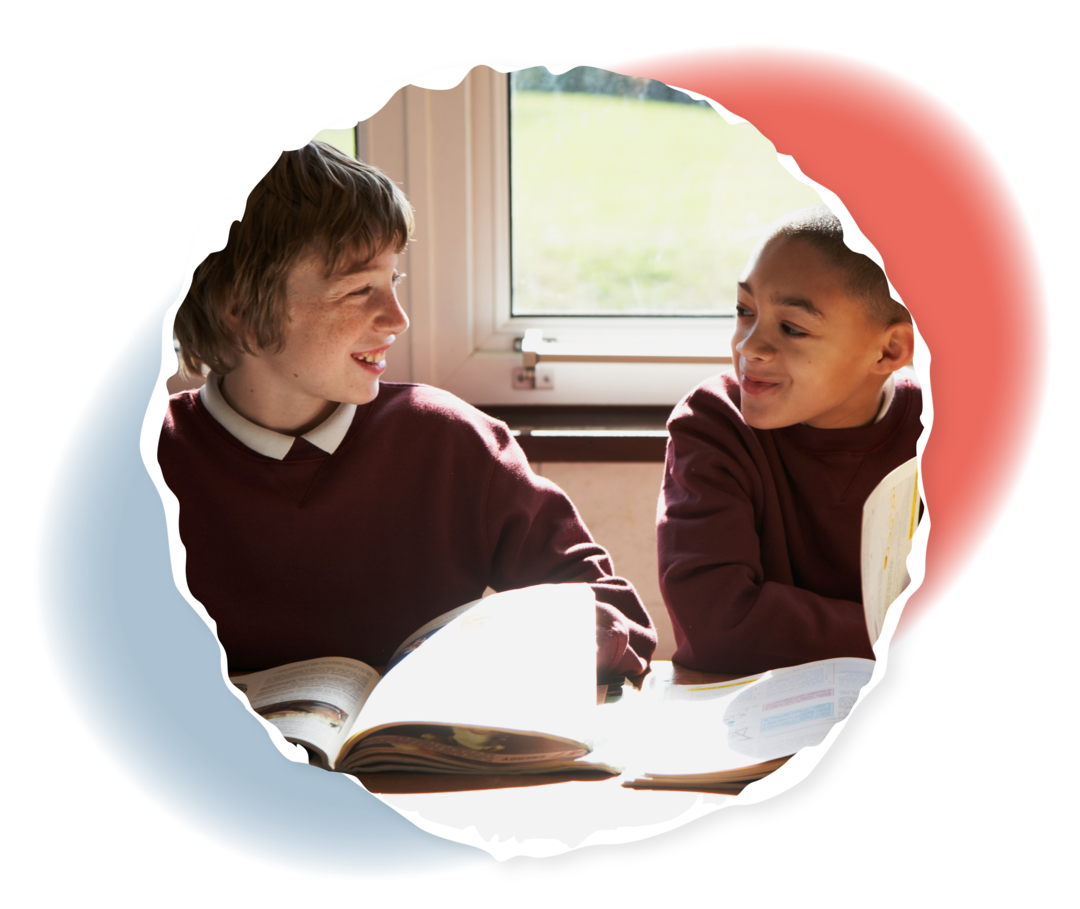 Zwei Jungen sitzen mit aufgeschlagenen Schulbüchern an einem Tisch und schauen sich lächelnd an.