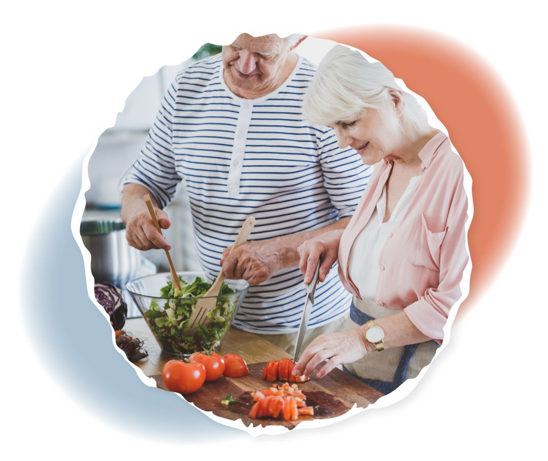 Eine Frau und ein Mann im Seniorenalter bereiten mit frischen Zutaten einen Salat zu: Die Frau schneidet Tomaten, der Mann mischt in einer Schüssel die Salatblätter. 